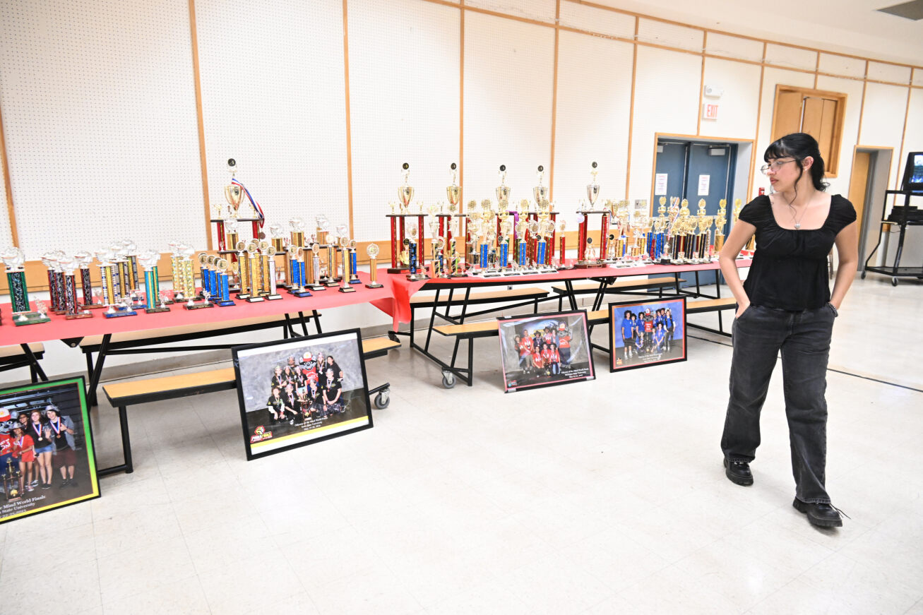 A girl with dark hair and a black shirt and pants looks back at photos and trophies from years past of Odyssey of the Mind